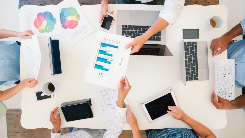 Masanın etrafında beş kişi, CRM verilerini inceleyerek birbirlerine sunum yapıyor, önlerinde laptoplar, tabletler ve tablo çıktıları bulunuyor.